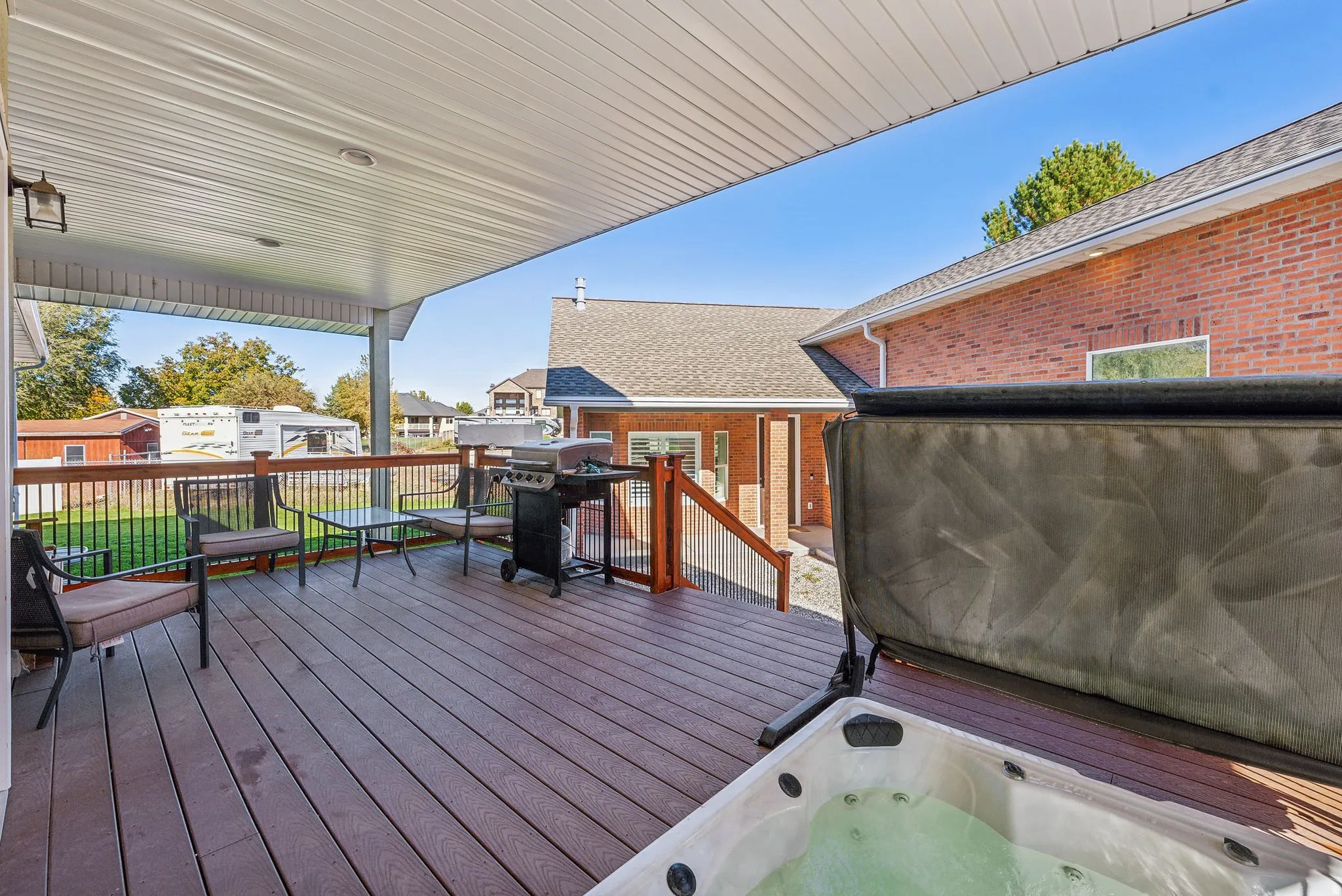 Spacious covered deck featuring composite decking, wood railing with black metal spindles, a built-in hot tub, and exterior lighting fixtures