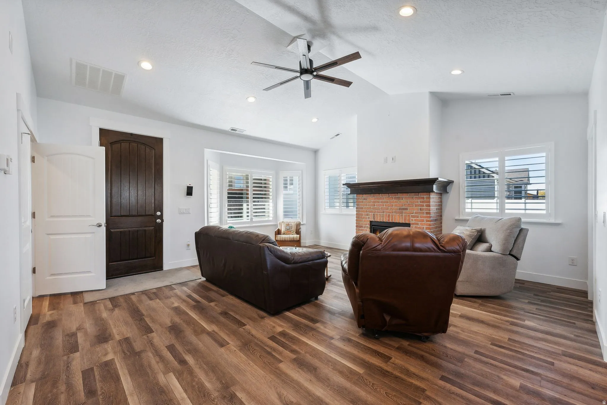 Custom built ADU/Spacious living area featuring wood-finish flooring, a brick fireplace with a dark wood mantel, and multiple windows with plantation shutters