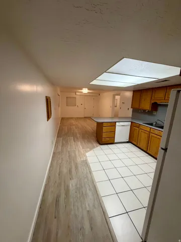 Open-concept kitchen and living area featuring wood-finish flooring in the main space, white ceramic tile in the kitchen, natural wood cabinetry, a built-in dishwasher, and a large overhead lighting panel