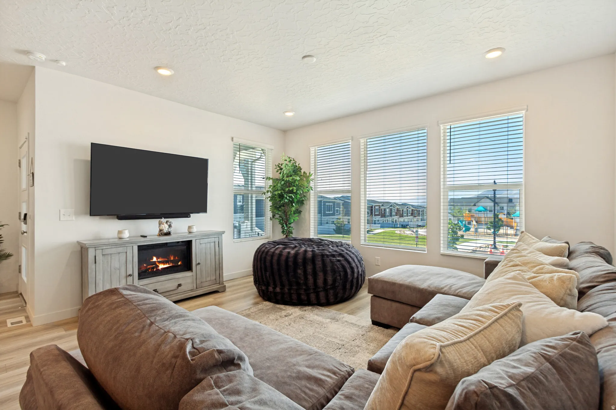 Living space featuring light wood-finish flooring, recessed ceiling lighting, and multiple windows with horizontal blinds