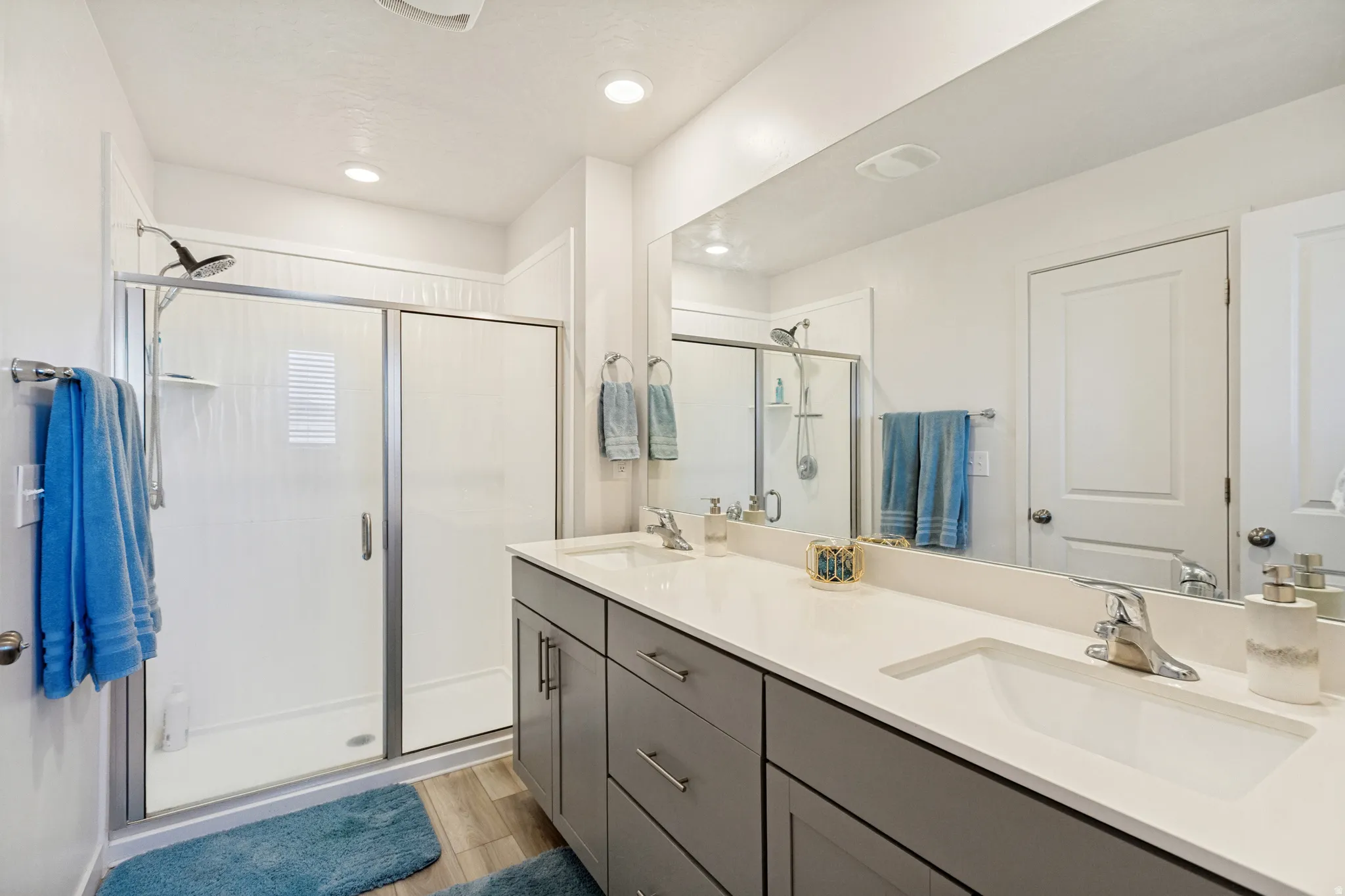 Bathroom featuring a dual vanity with grey cabinetry and light-colored countertops, a large frameless mirror, a shower with a sliding glass door, and wood-finish flooring