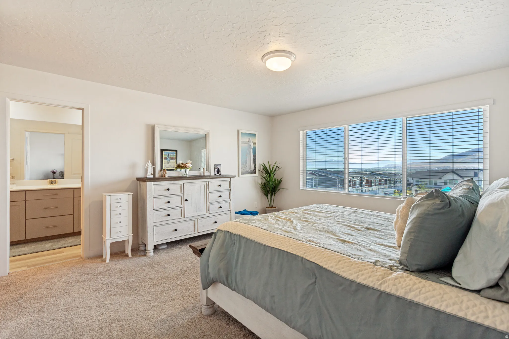 Carpeted bedroom featuring a large window with blinds, ceiling-mounted light fixture, and an open doorway to a bathroom with a vanity