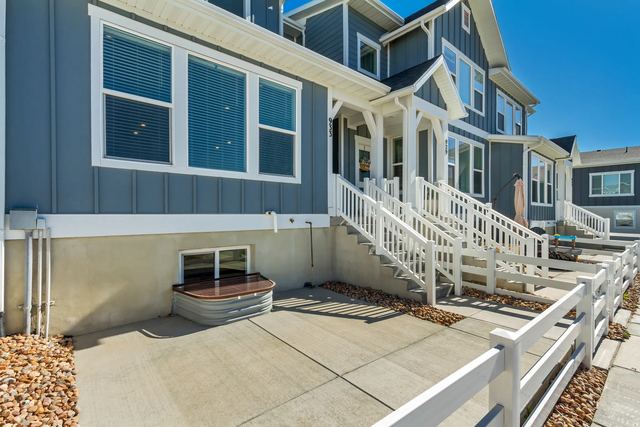 Exterior featuring blue vertical siding, white trim, multiple windows with blinds, a concrete patio area, and a white picket fence