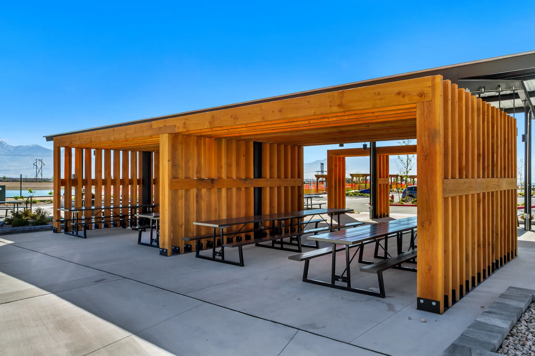 Outdoor wood-finish pavilion featuring multiple picnic tables with metal frames and seating, a concrete patio, and slatted wood-finish walls