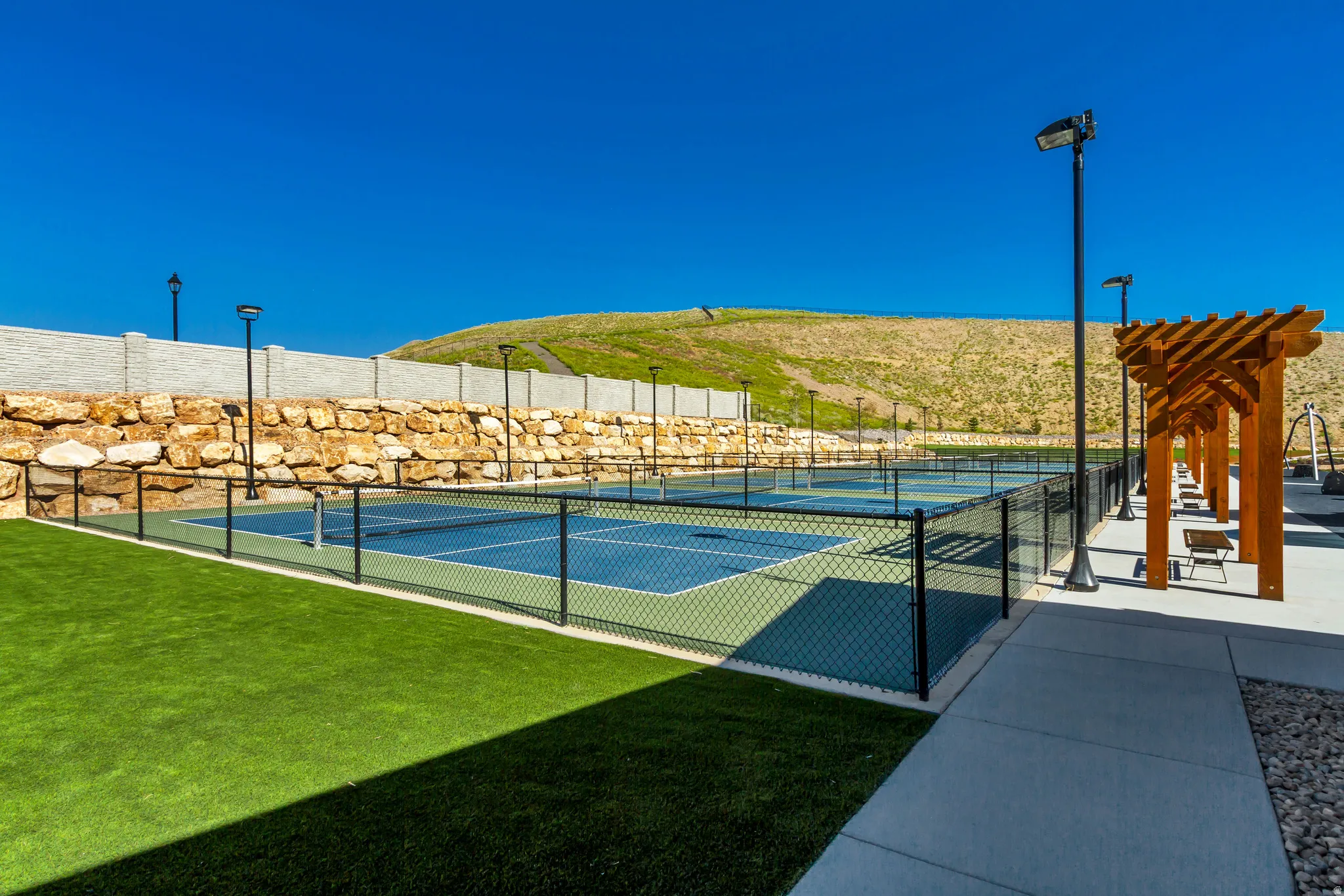 Outdoor pickleball courts featuring a black chain-link fence, surrounding artificial turf, and adjacent concrete pathways