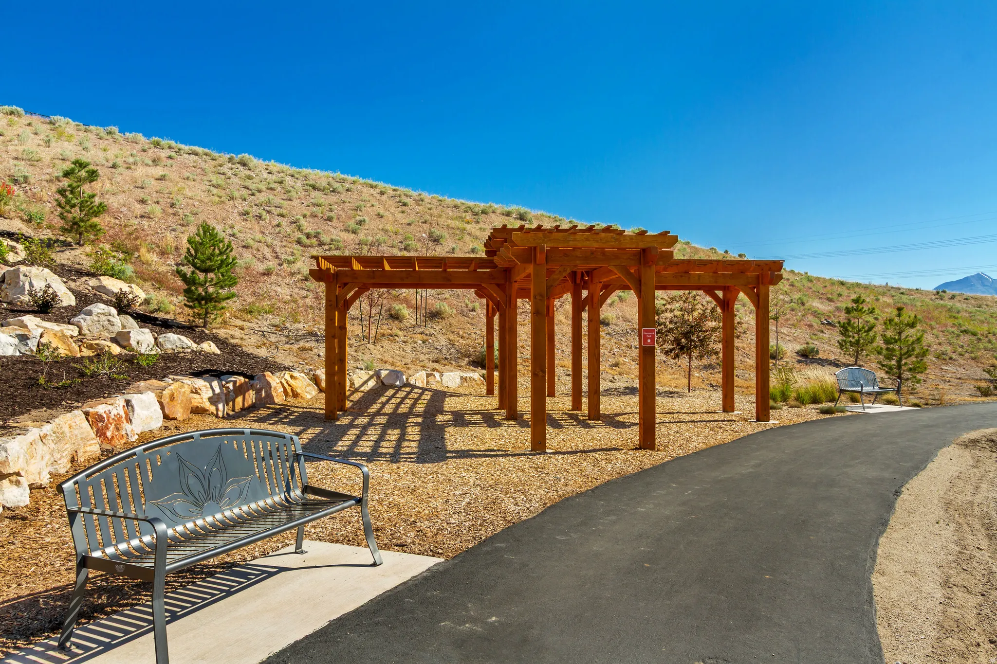 Wood pergola structure with a chip bark ground cover, bordered by a paved pathway and a landscaped rock garden