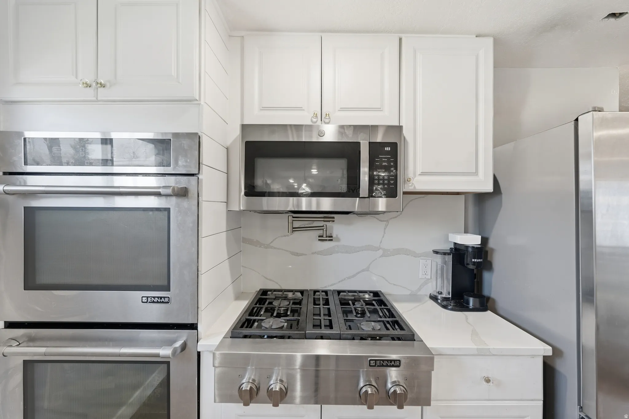 Kitchen featuring stainless steel double wall ovens, a stainless steel microwave, a gas range with a pot filler, white shaker-style cabinetry, and white countertops with a subtle gray veining