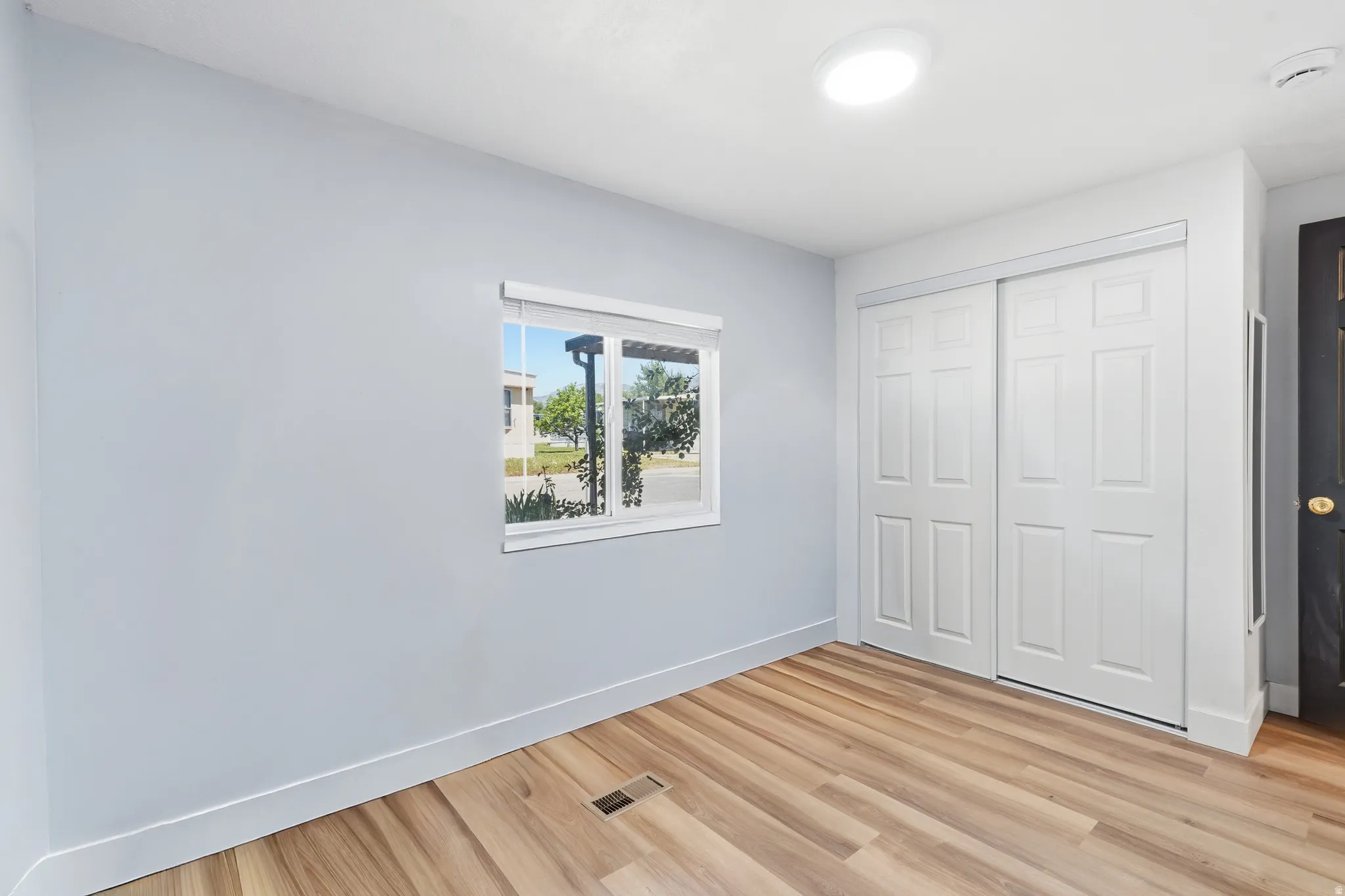 Room featuring wood-finish flooring, a window with a roller shade, and white paneled sliding closet doors