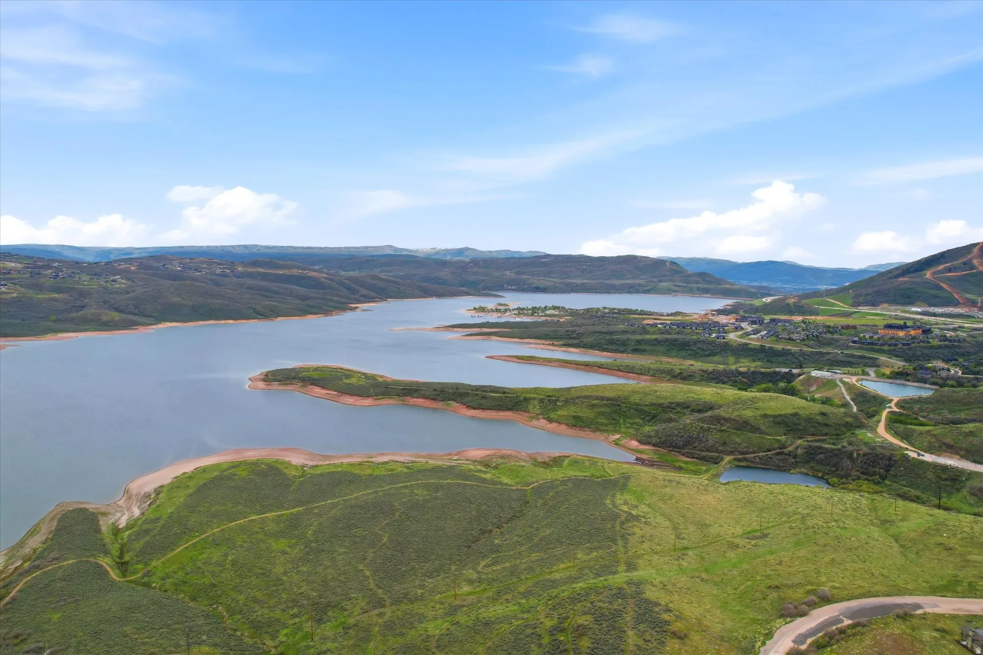 Expansive lake surrounded by rolling green hills and distant mountains under a blue sky with scattered clouds