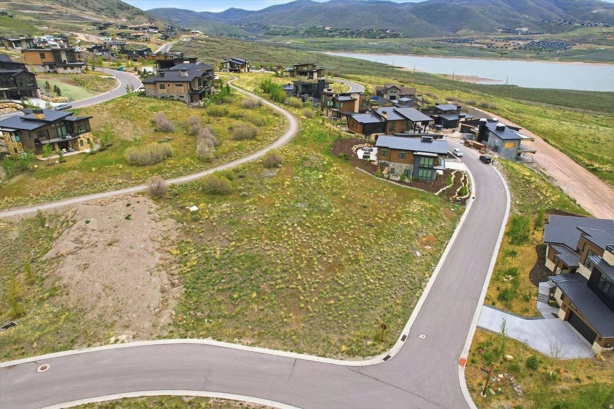 Expansive undeveloped hillside lot featuring a paved road frontage, surrounding modern residences, and distant lake and mountain scenery