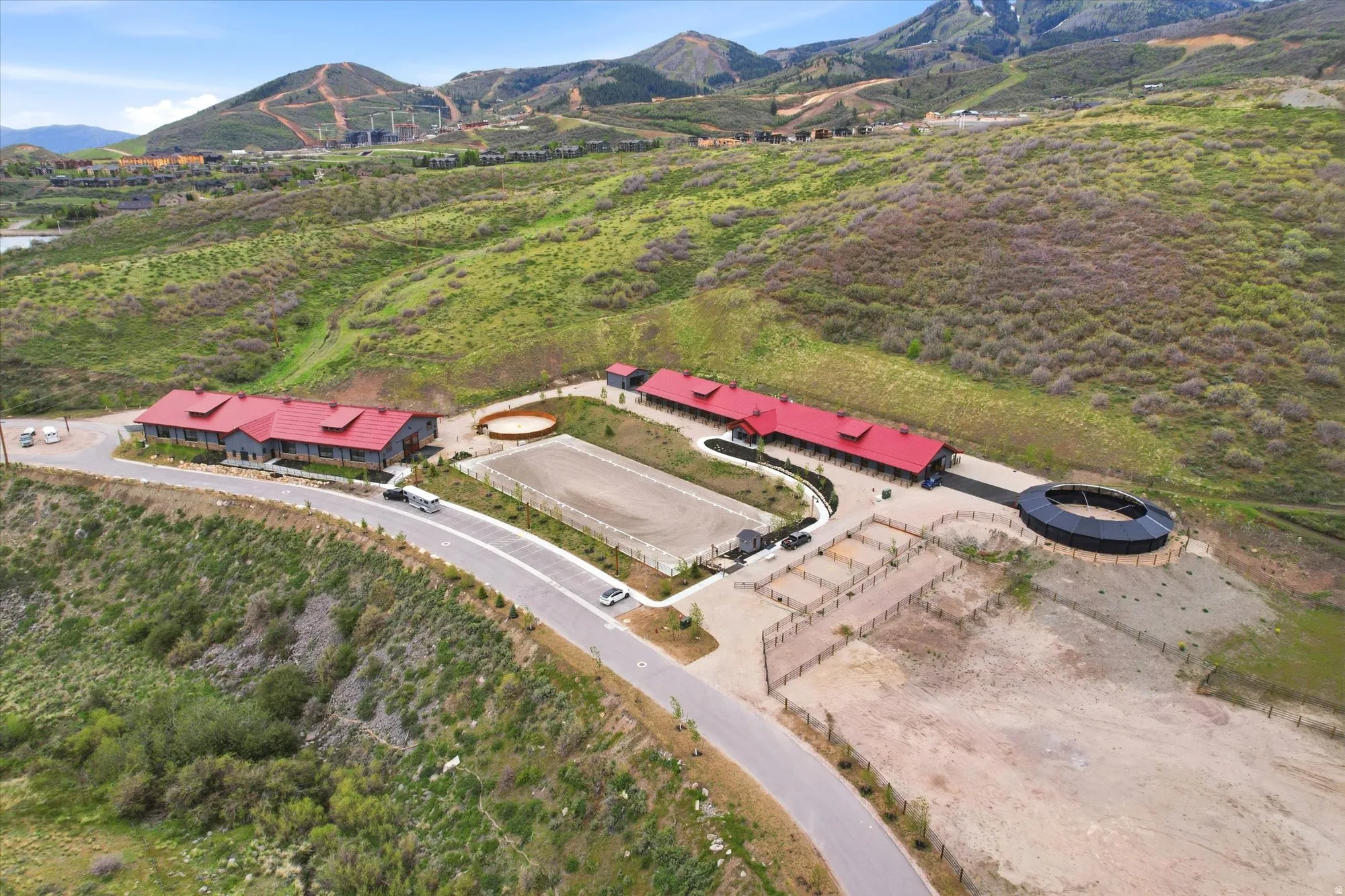Expansive equestrian facility featuring multiple buildings with red roofs, a large outdoor arena, several fenced paddocks, and a round pen