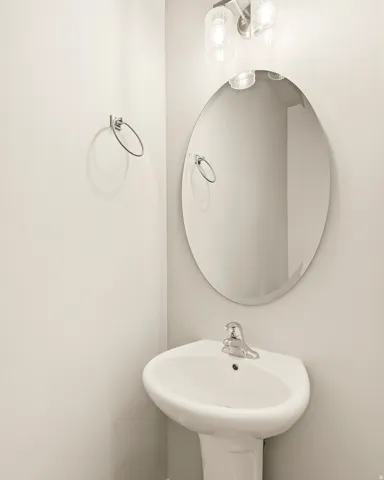 White pedestal sink with a chrome faucet, illuminated by a three-light vanity fixture, complemented by an oval mirror and a wall-mounted towel ring