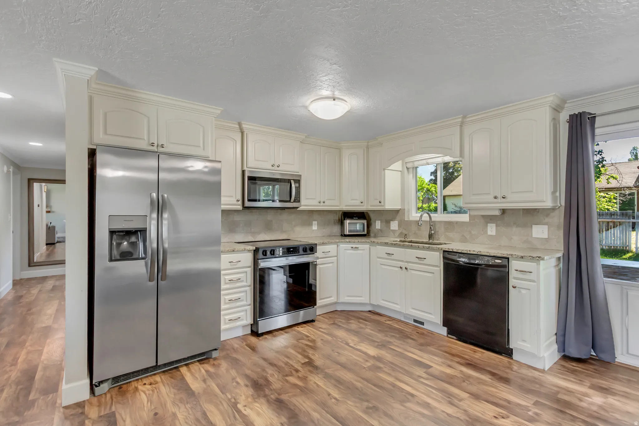 Spacious kitchen featuring wood-finish flooring, white cabinetry with crown molding, stainless steel appliances, granite countertops, and a tiled backsplash
