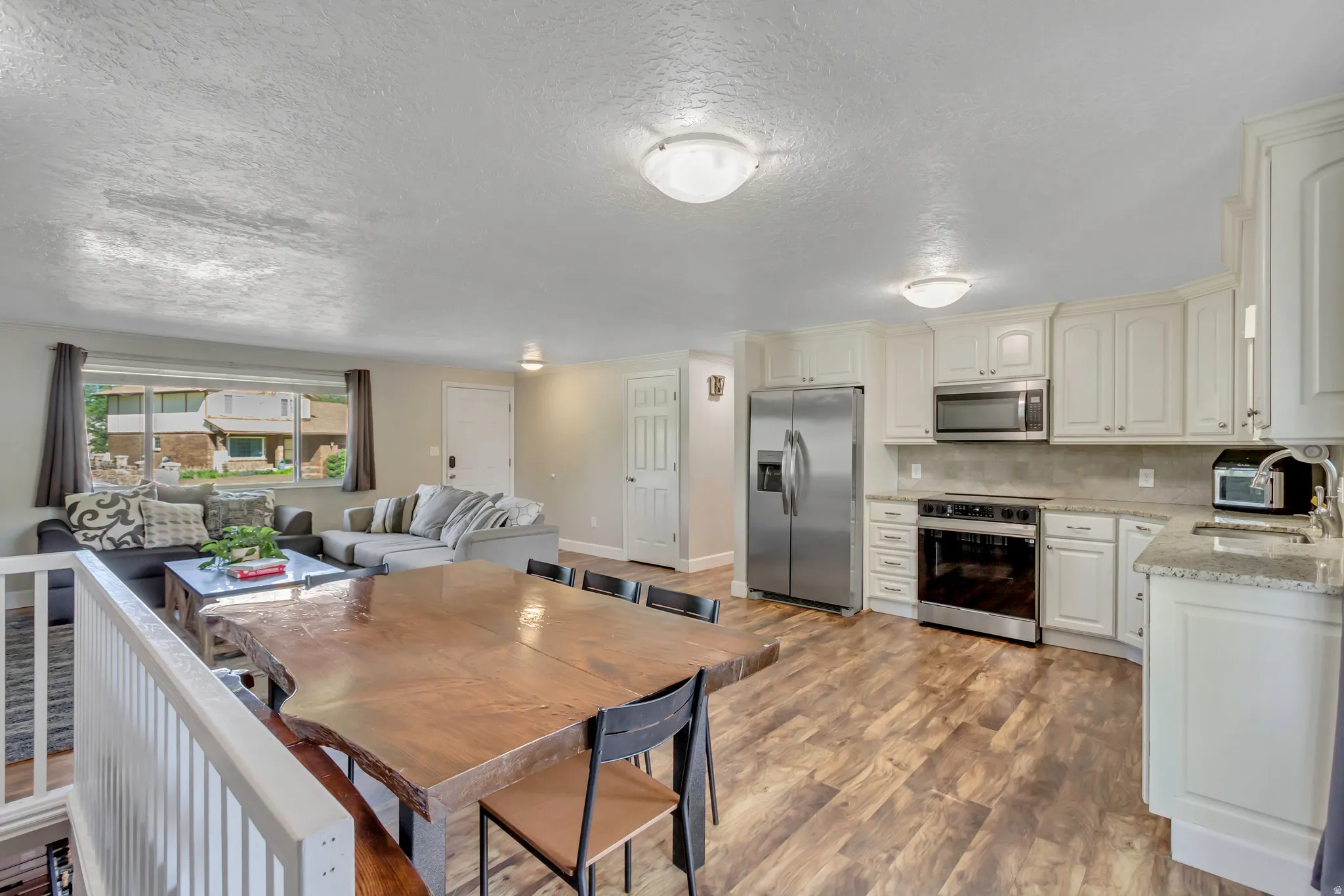 Open-concept living space featuring wood-finish flooring, white cabinetry, granite-style countertops, stainless steel appliances, and a large window