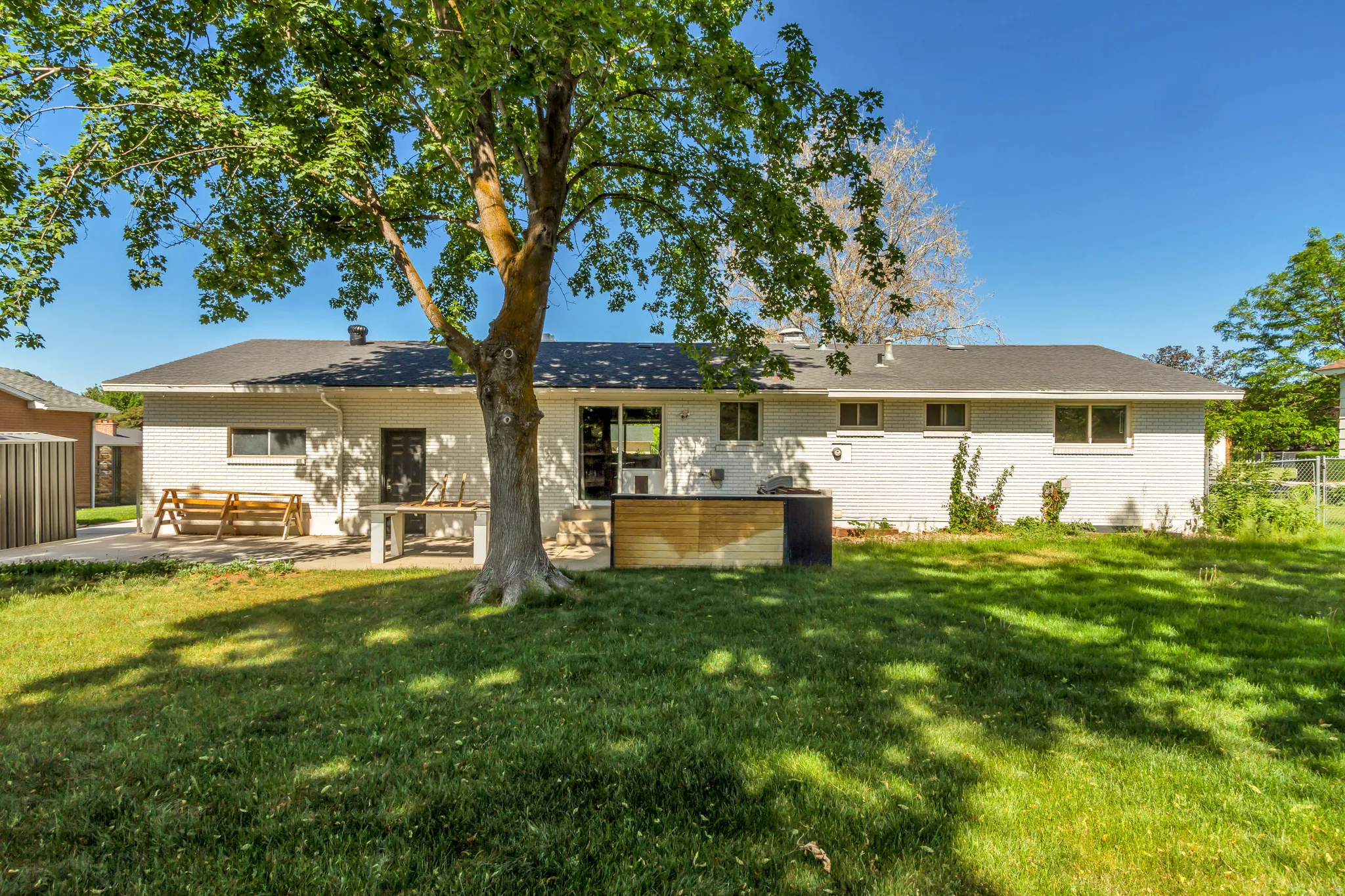Expansive backyard featuring a mature tree providing natural shade, a white brick exterior, a black shingle roof, a concrete patio area, and a wood-slat enclosure
