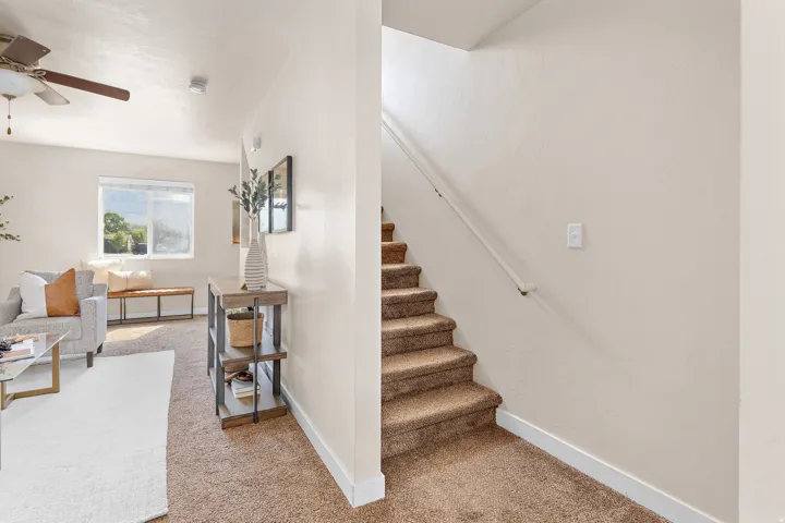 Carpeted staircase with white handrail alongside a living area featuring a ceiling fan, large window, and light-toned walls