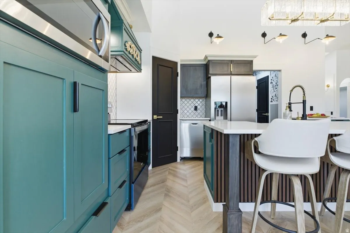 Kitchen featuring teal cabinetry with black hardware, stainless steel appliances, a white island with slatted wood detailing, herringbone wood-finish flooring, and a patterned tile backsplash