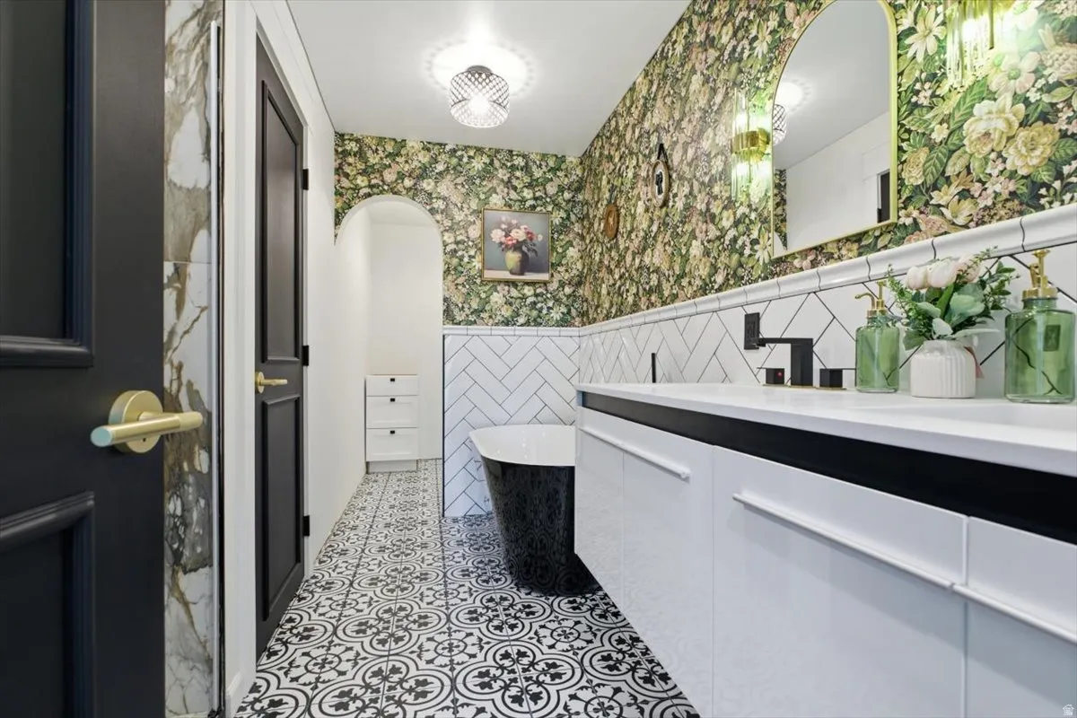 Bathroom featuring a freestanding black tub, patterned floor tiles, white subway tile wainscoting, a double vanity with white cabinetry and integrated sinks, and an arched mirror