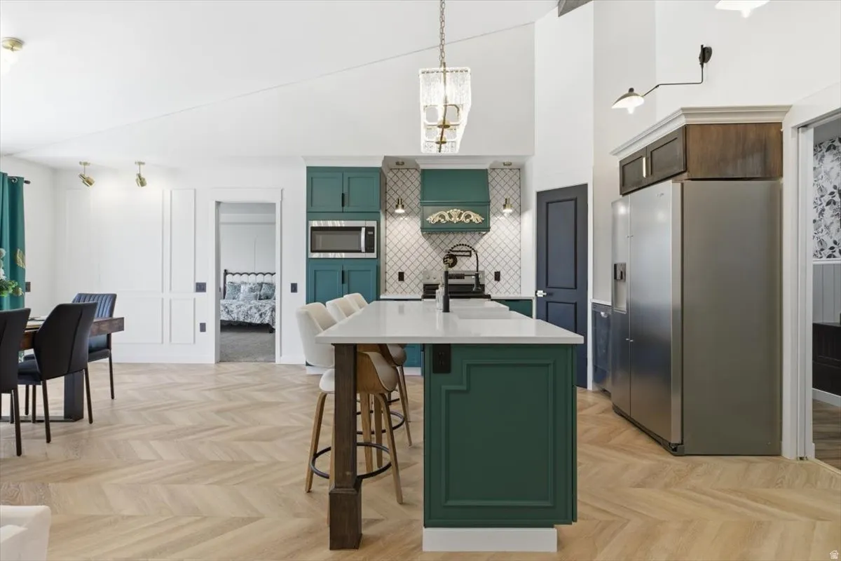 Open-concept kitchen featuring an island with seating, teal cabinetry, a diagonal tile backsplash, stainless steel appliances, and herringbone-patterned wood-finish flooring