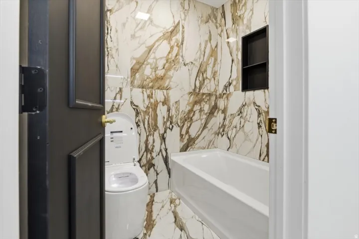 Full bathroom featuring large format veined wall and floor tiles, a white soaking tub, and a built-in shower niche