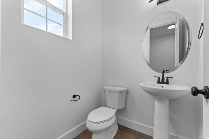 Powder room featuring a pedestal sink with a matte black faucet, a round mirror, and wood-finish flooring