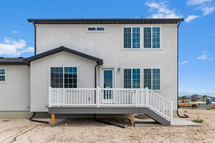 Rear exterior featuring light-toned stucco, multiple windows with grids, a ground-level deck with white railings, a patio door, and a gray-trimmed roofline