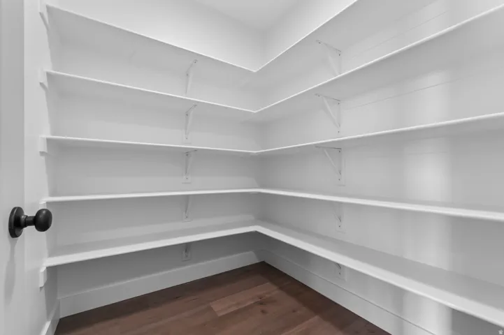 Spacious pantry featuring multiple tiers of white shelving, wood-finish flooring, and a matte black door knob