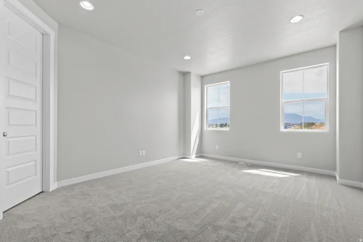 Bright room featuring two large windows, recessed ceiling lighting, light gray carpeting, and a white paneled door with brushed nickel hardware