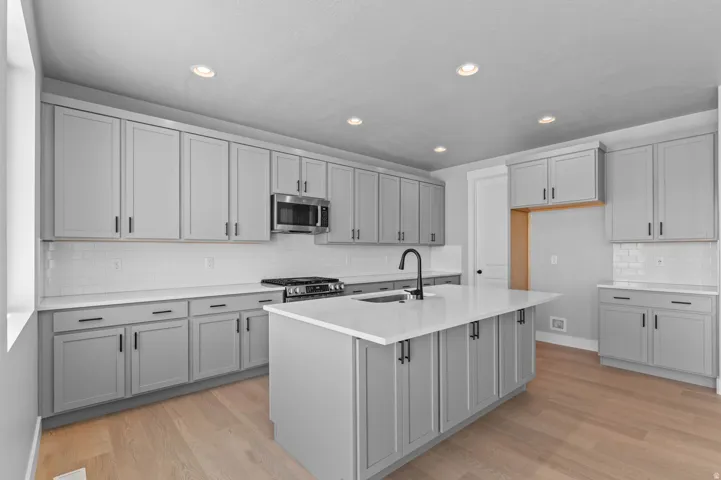 Kitchen featuring a central island with an integrated sink, light gray shaker-style cabinetry with matte black hardware, white subway tile backsplash, stainless steel appliances, and light wood-finish flooring