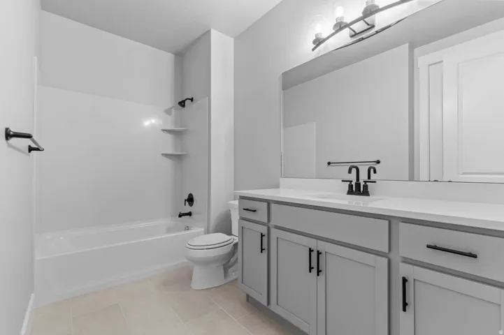Full bathroom featuring a white tub/shower combination with integrated shelving, a single vanity with a white countertop, and light gray cabinetry with matte black hardware
