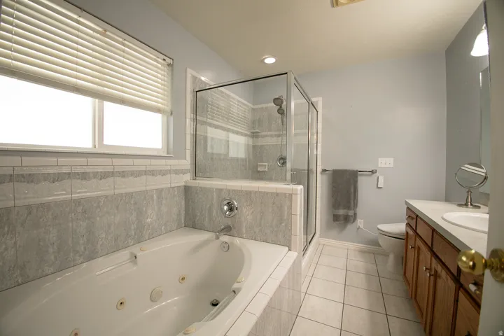 Primary bathroom with Jetted tub with tiled surround, glass-enclosed shower, white tile flooring, double-pane window with blinds, and a wood-finish vanity with countertop sink