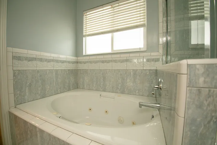 Corner jetted tub with tiled surround, light blue painted walls, and window with blinds