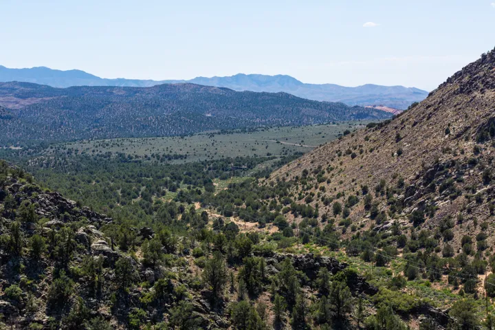 Expansive mountain landscape featuring varied terrain, distant mountain ranges, a verdant valley, and a mix of sparse and dense vegetation
