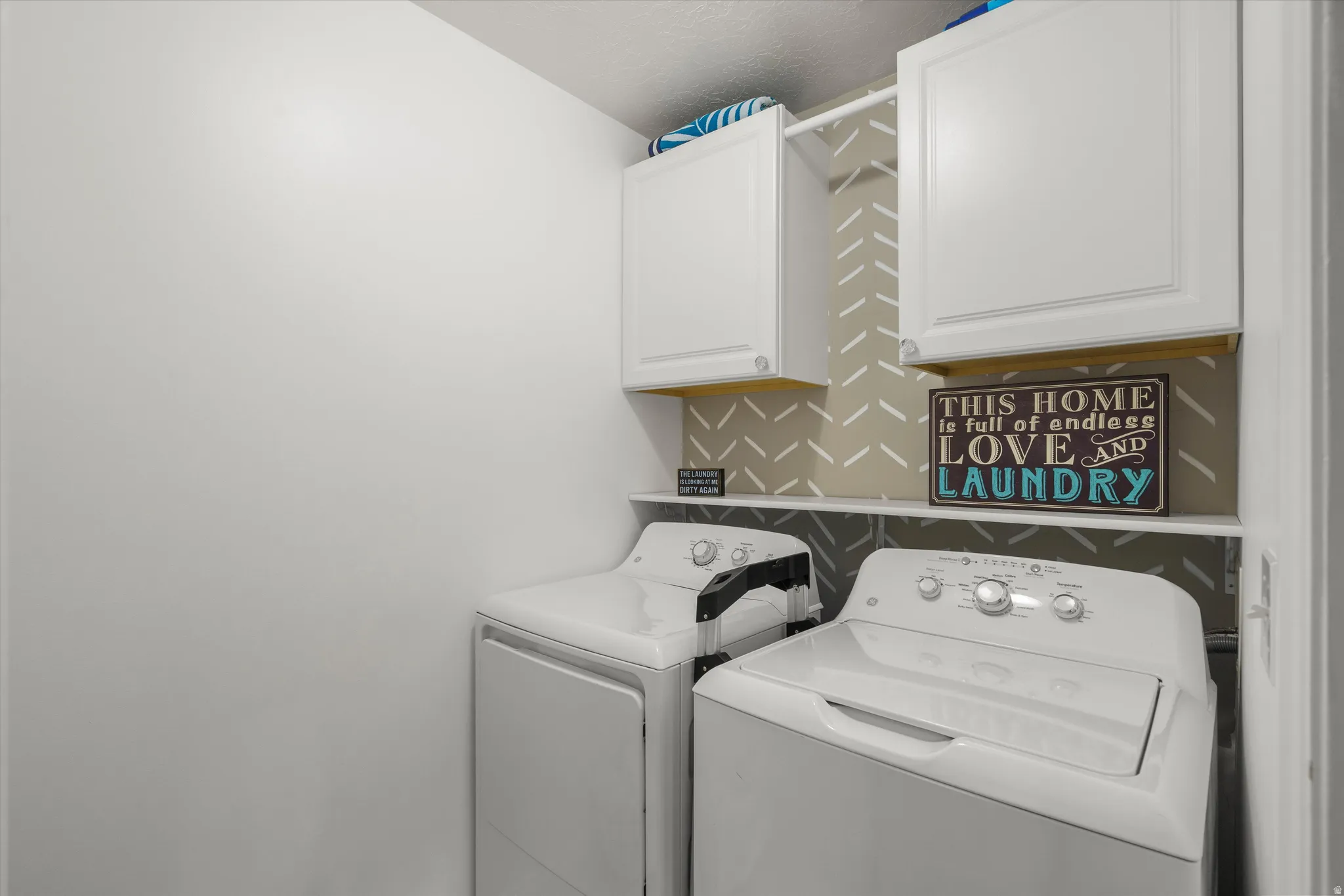 Dedicated laundry area featuring a patterned accent wall, white upper cabinetry, and a white storage shelf