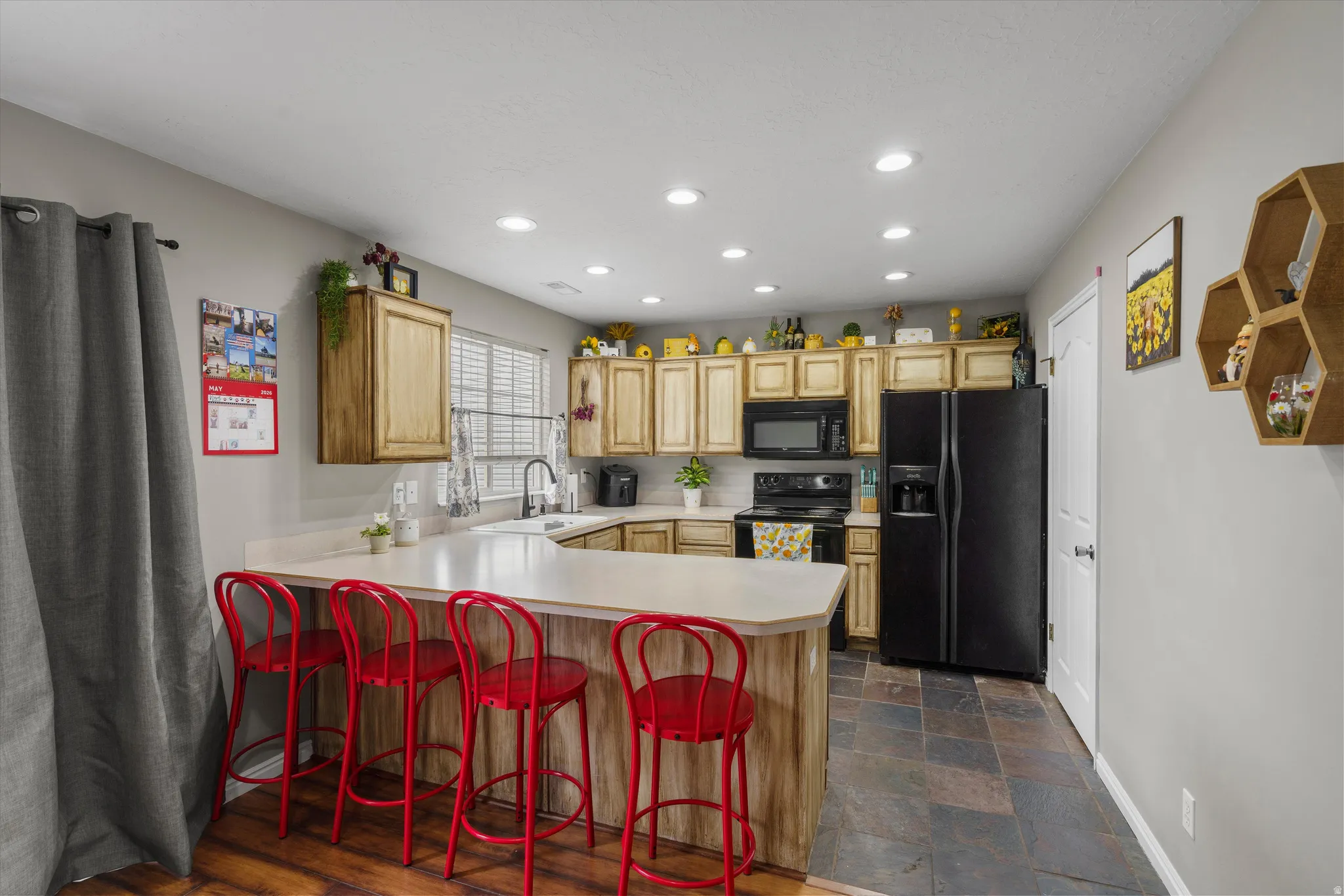 Kitchen featuring a U-shaped island with a light-toned countertop, wood-finish cabinetry, black appliances, recessed lighting, and slate-style floor tiling. Oversized pantry