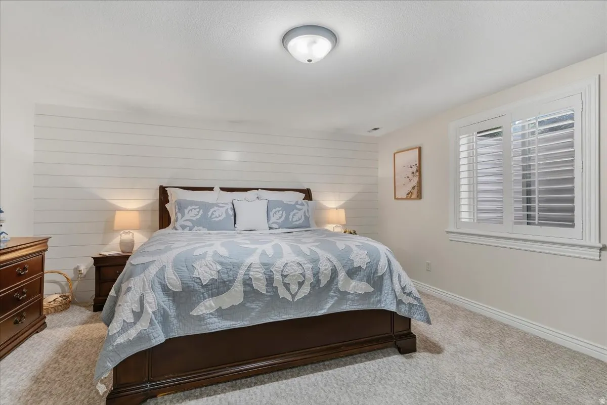 Bedroom featuring a shiplap accent wall, white plantation shutters, neutral wall paint, ceiling-mounted light fixture, and plush carpeting