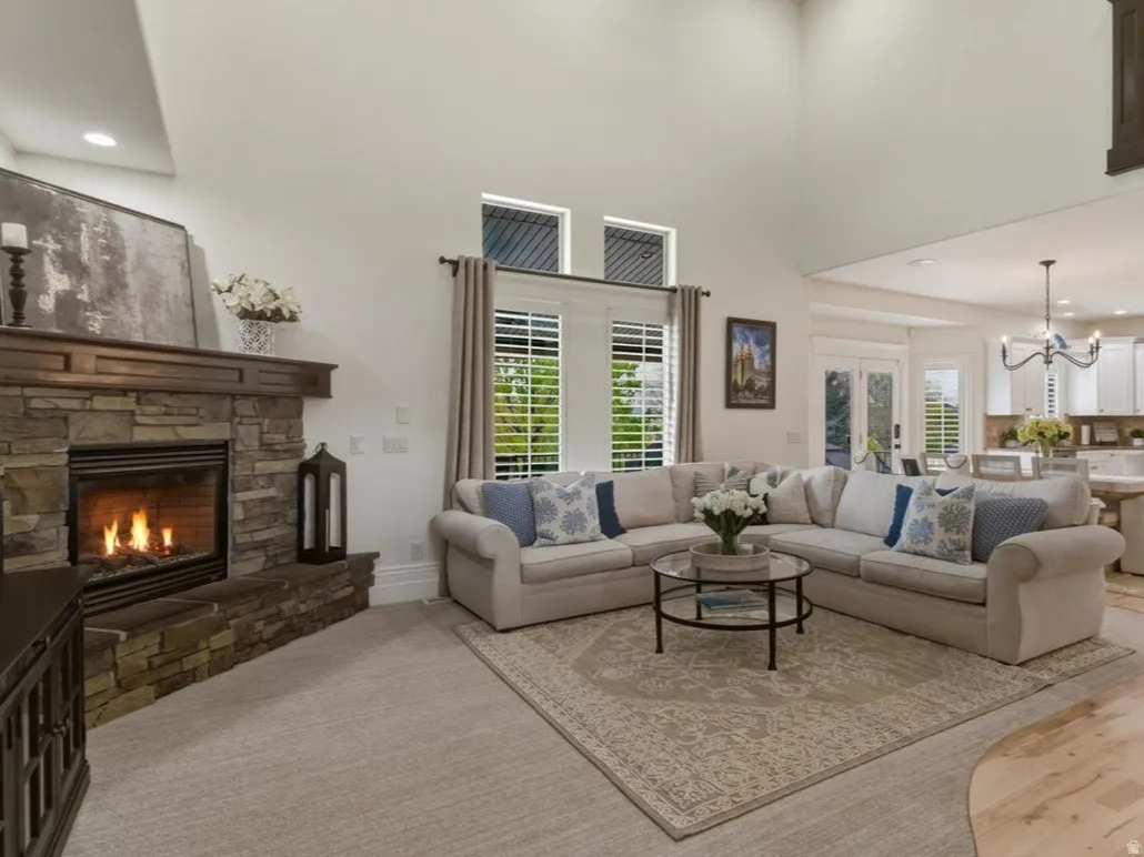Spacious living area featuring a stacked stone fireplace with a wood mantel, high ceilings, large windows with white louvered shutters, and a partial view of a kitchen with light wood flooring