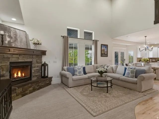 Spacious living area featuring a stacked stone fireplace with a wood mantel, high ceilings, large windows with white louvered shutters, and a partial view of a kitchen with light wood flooring