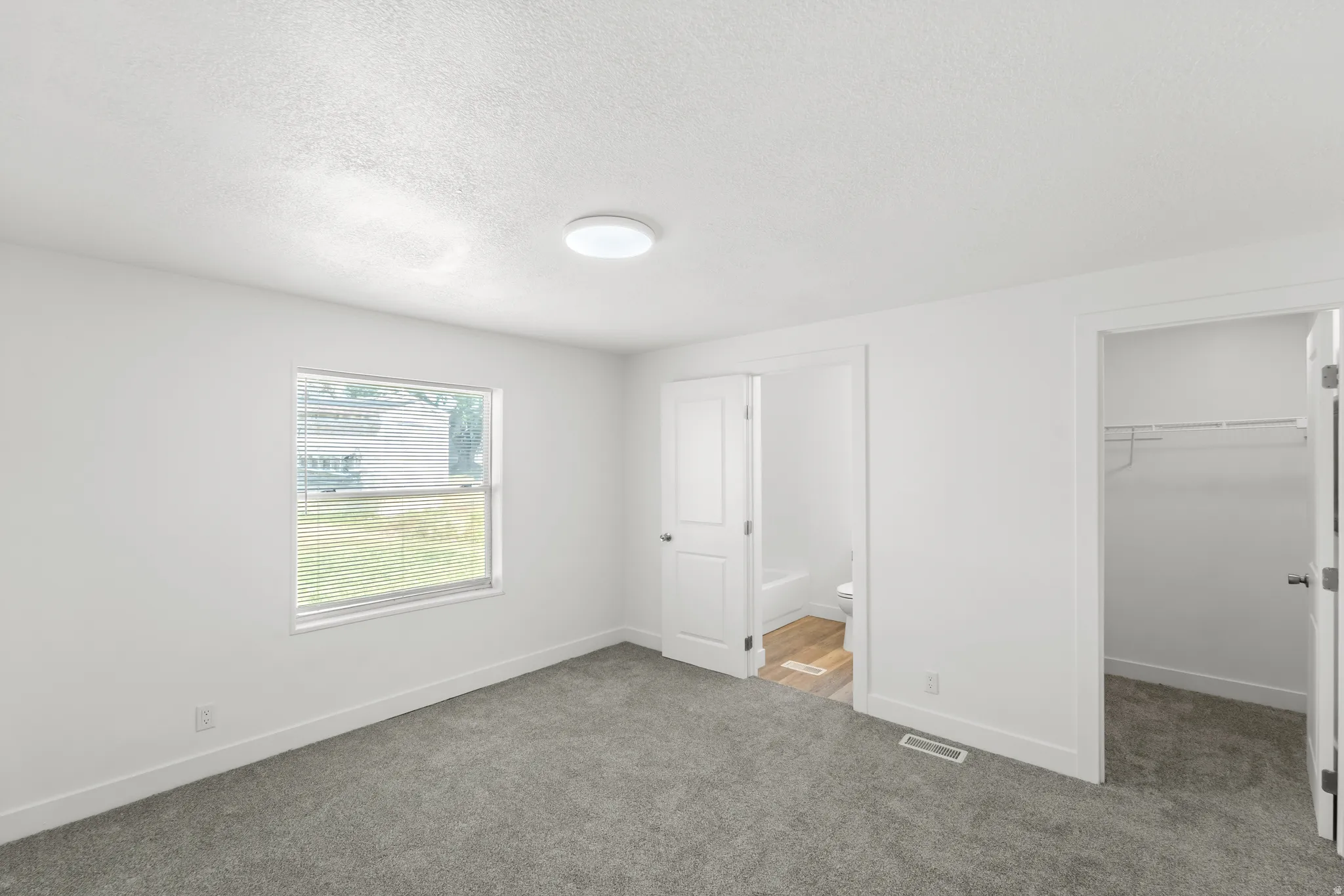 Bright room featuring a window with blinds, a ceiling-mounted light fixture, plush gray carpeting, white baseboards, and a bi-fold closet door with wire shelving