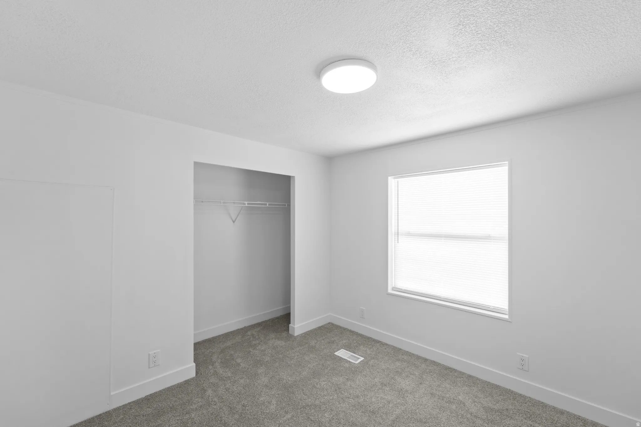 Carpeted room featuring a single window with blinds, a built-in closet with wire shelving, a ceiling-mounted light fixture, and white baseboards