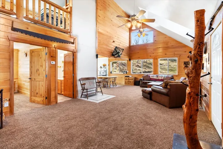 Vaulted ceiling great room featuring natural wood paneling, multiple windows, a ceiling fan with integrated lighting, and a carpeted floor