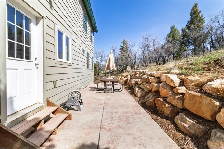 Exterior patio featuring a concrete surface, a stacked stone retaining wall, and natural landscaping with mature trees