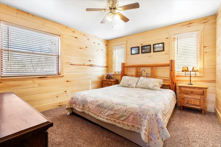 Bedroom featuring extensive wood paneling, two windows with blinds, and a built-in ceiling fan with lighting