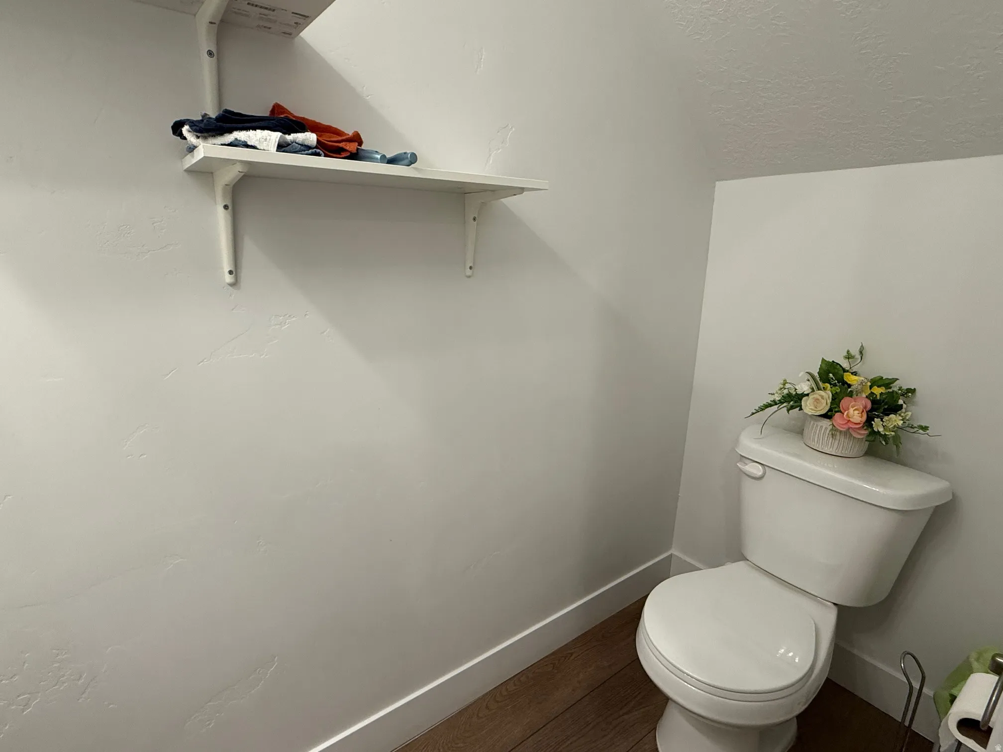 White toilet with a built-in shelving unit, white walls, and wood-finish flooring