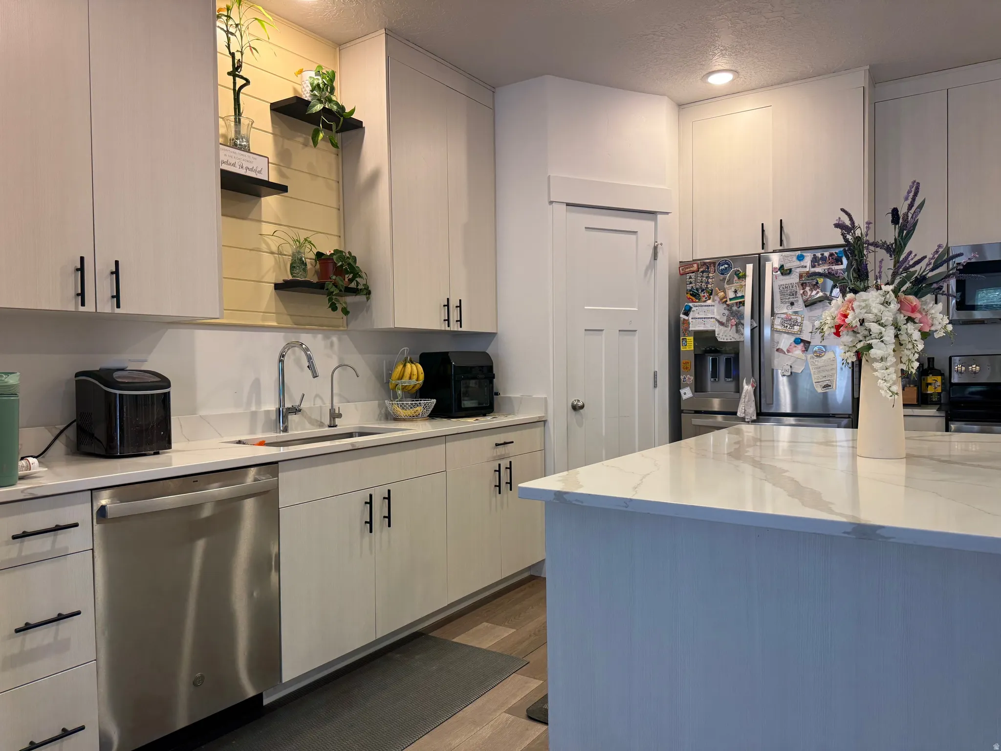 Contemporary kitchen featuring light-toned cabinetry with matte black hardware, stainless steel appliances, white countertops, and a contrasting wood-paneled backsplash accent