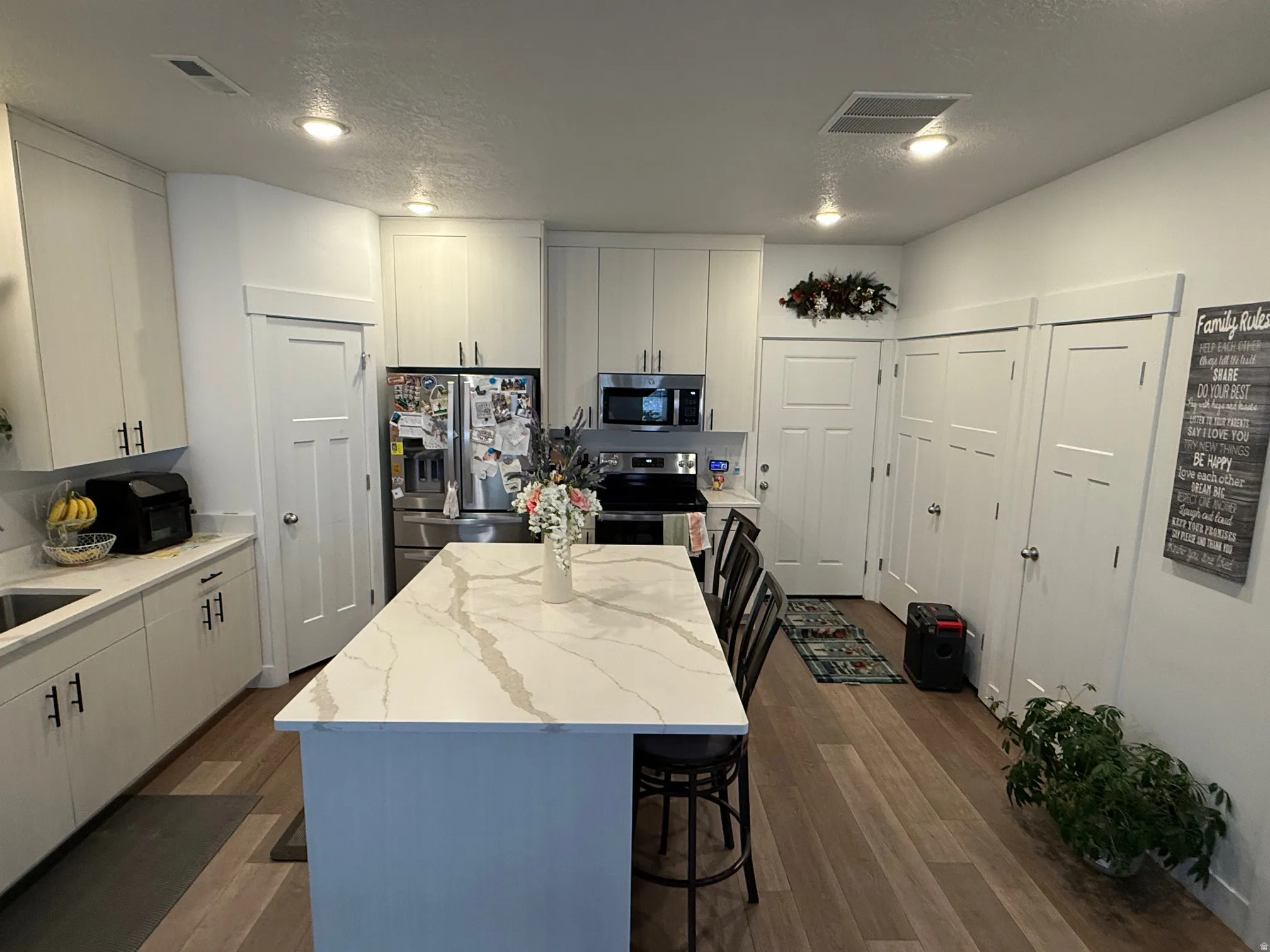 Modern kitchen featuring a large central island with a waterfall-edge countertop, light wood-finish flooring, recessed lighting, white cabinetry, and stainless steel appliances