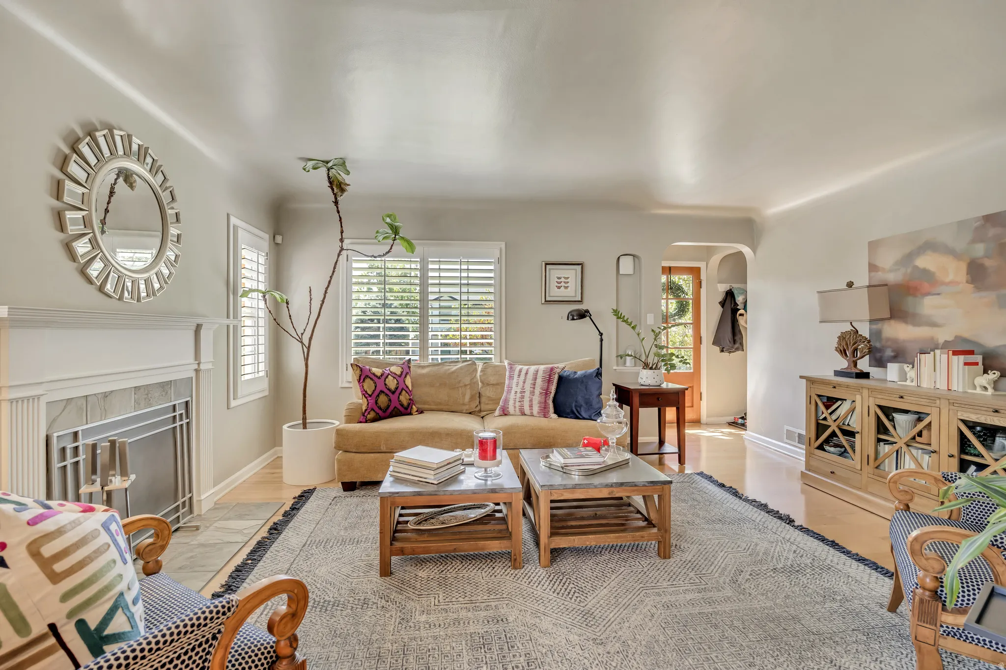 Living area featuring light wood-finish flooring, a decorative fireplace with a white mantel and tile surround, white plantation shutters, and a recessed archway