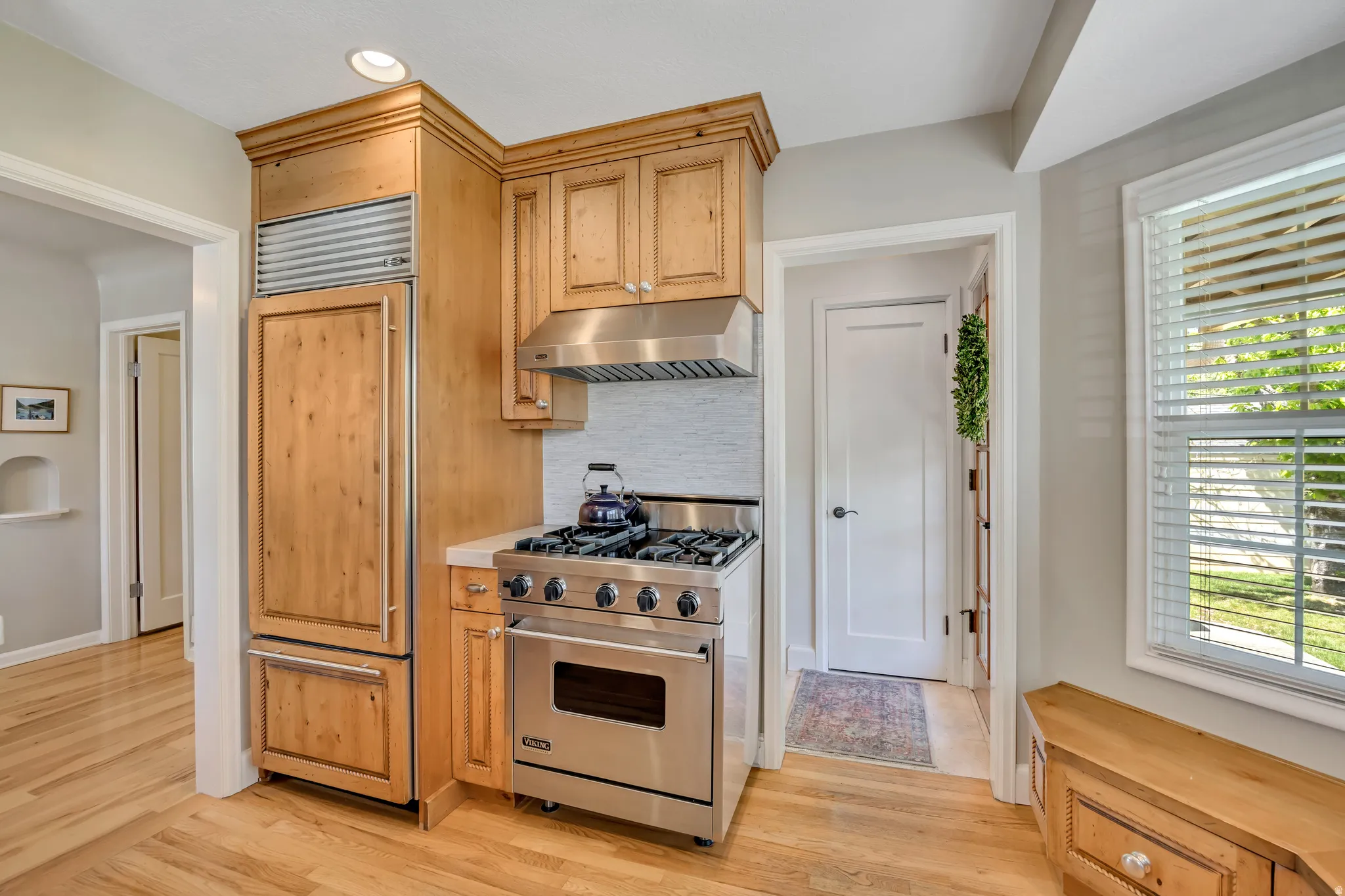 Kitchen featuring stainless steel appliances, custom paneled refrigerator, natural wood cabinetry, light tile backsplash, and light wood-finish flooring