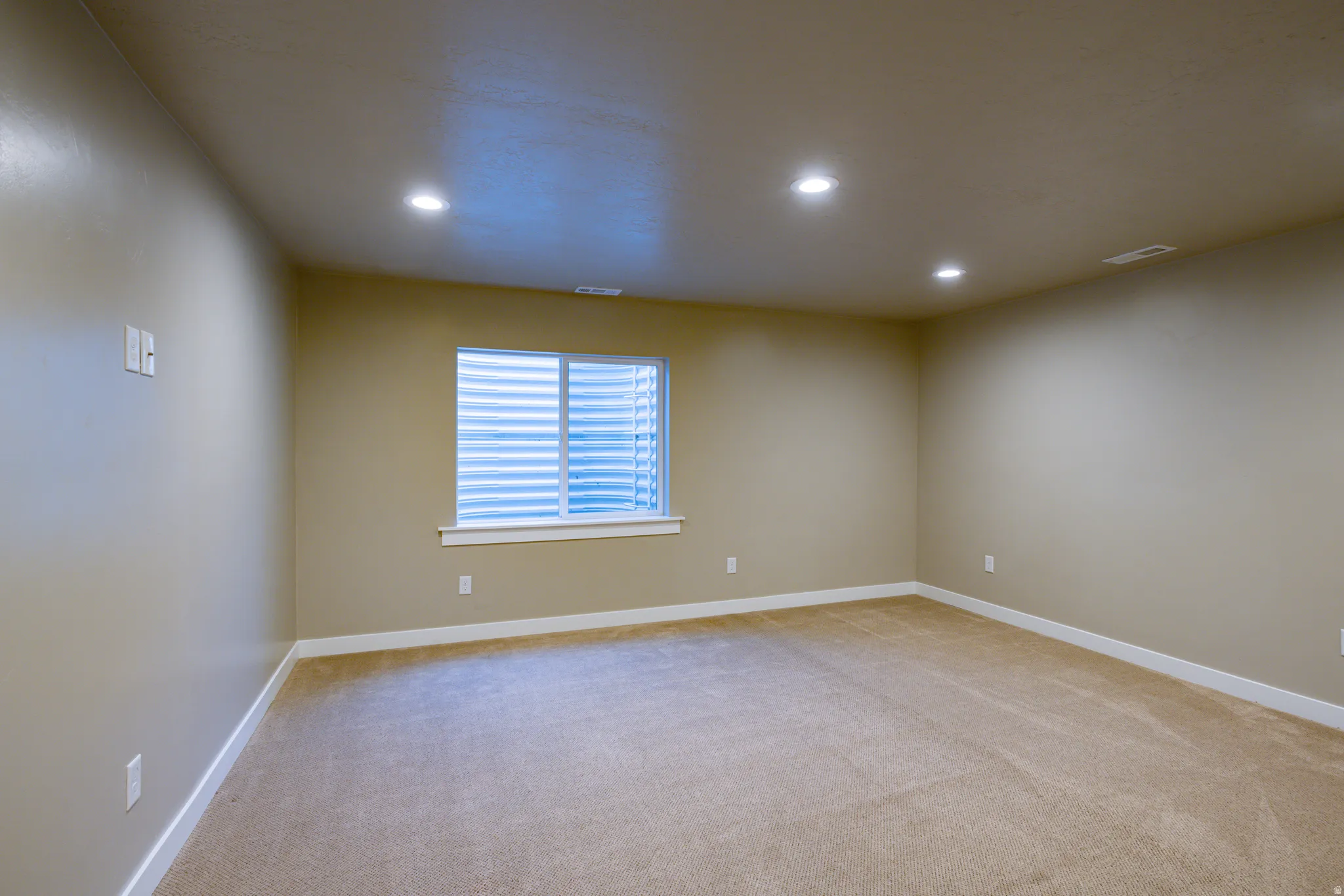 Spacious room featuring neutral carpeting, recessed ceiling lighting, and a white-trimmed window with horizontal blinds