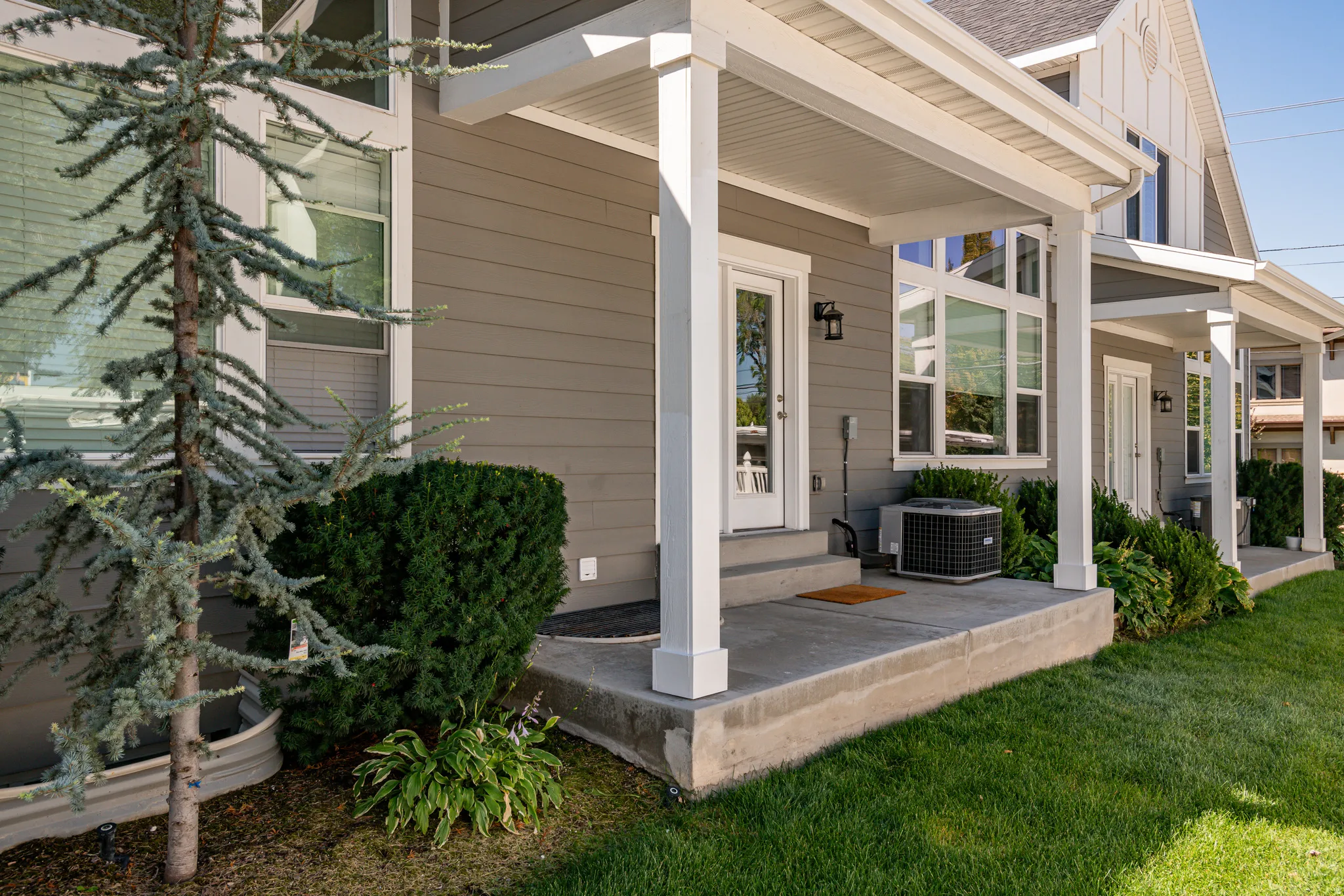 Exterior featuring gray siding with white trim, a concrete porch, multiple windows, and manicured landscaping with a small tree